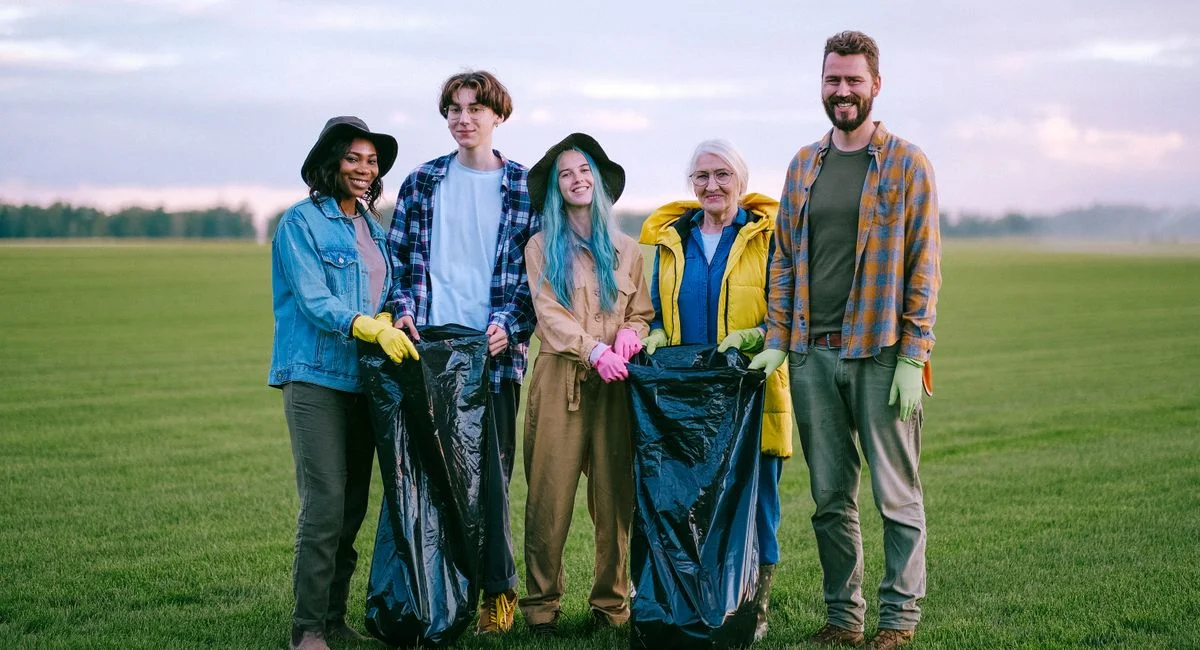 A group of diverse volunteers holding garbage bags while cleaning a green field outdoors.