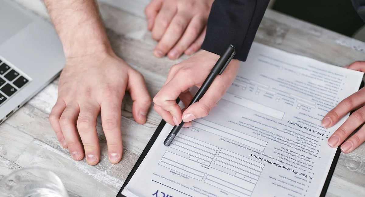 Close-up image of two people signing an insurance policy document on a wooden desk.