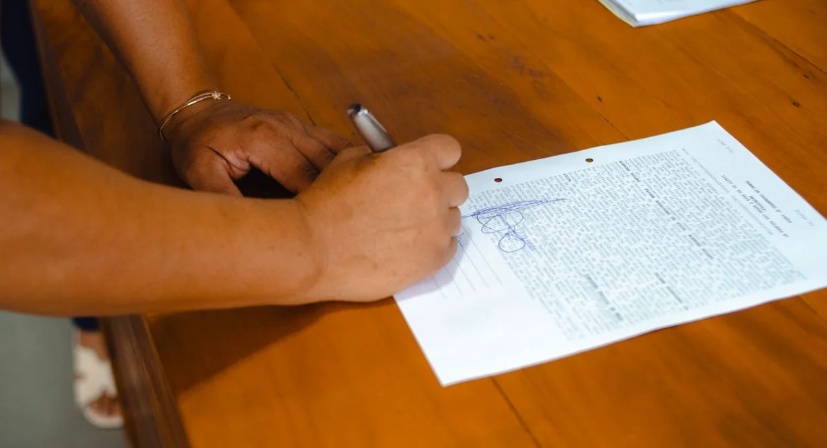 Close-up of hands signing a legal document on a wooden table indoors.