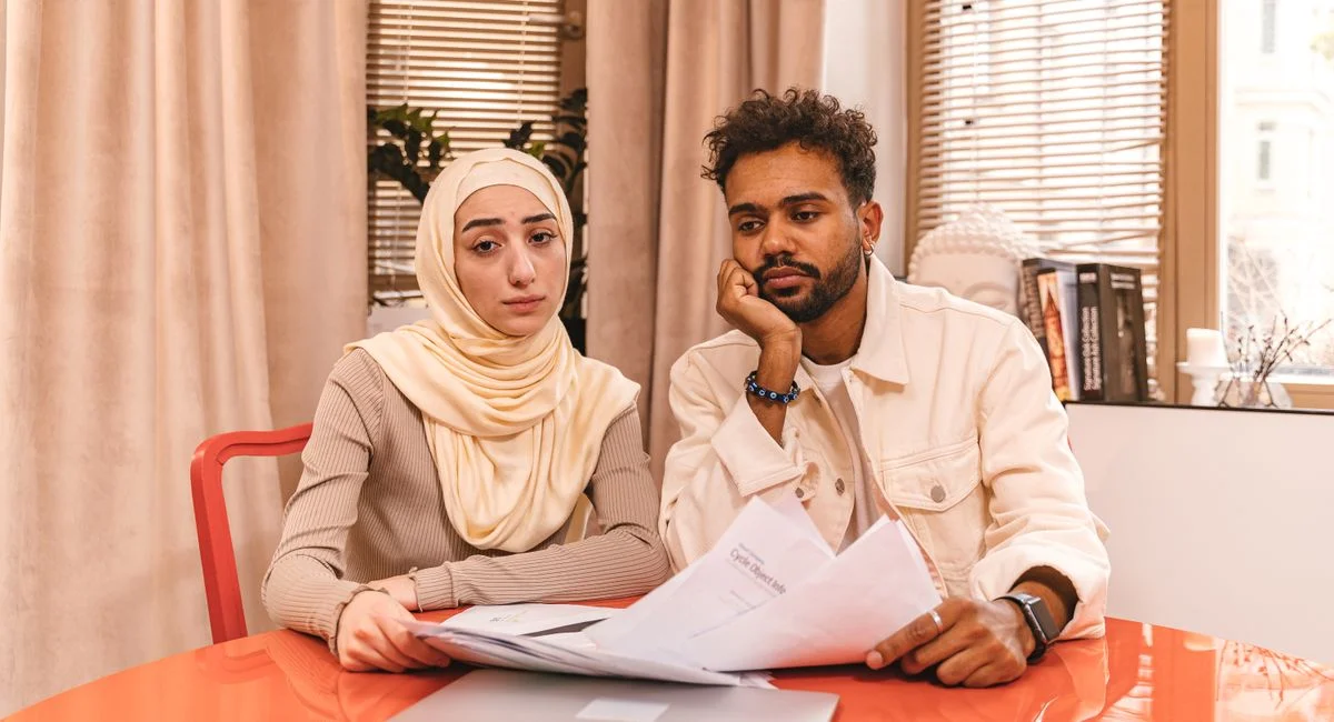 A couple in casual attire sits indoors at a table reviewing financial documents with a laptop.