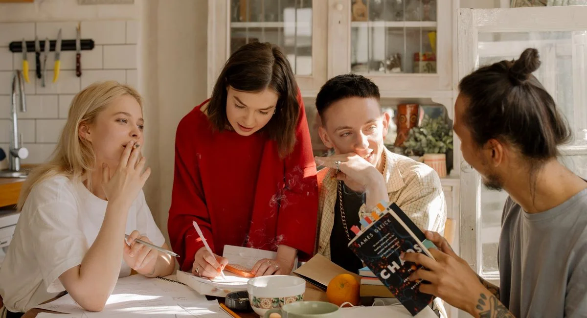 Group of young adults studying together at a kitchen table filled with books and notes.