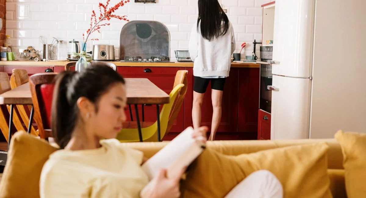Two women relax at home. One reads on the sofa while the other is at the kitchen counter.