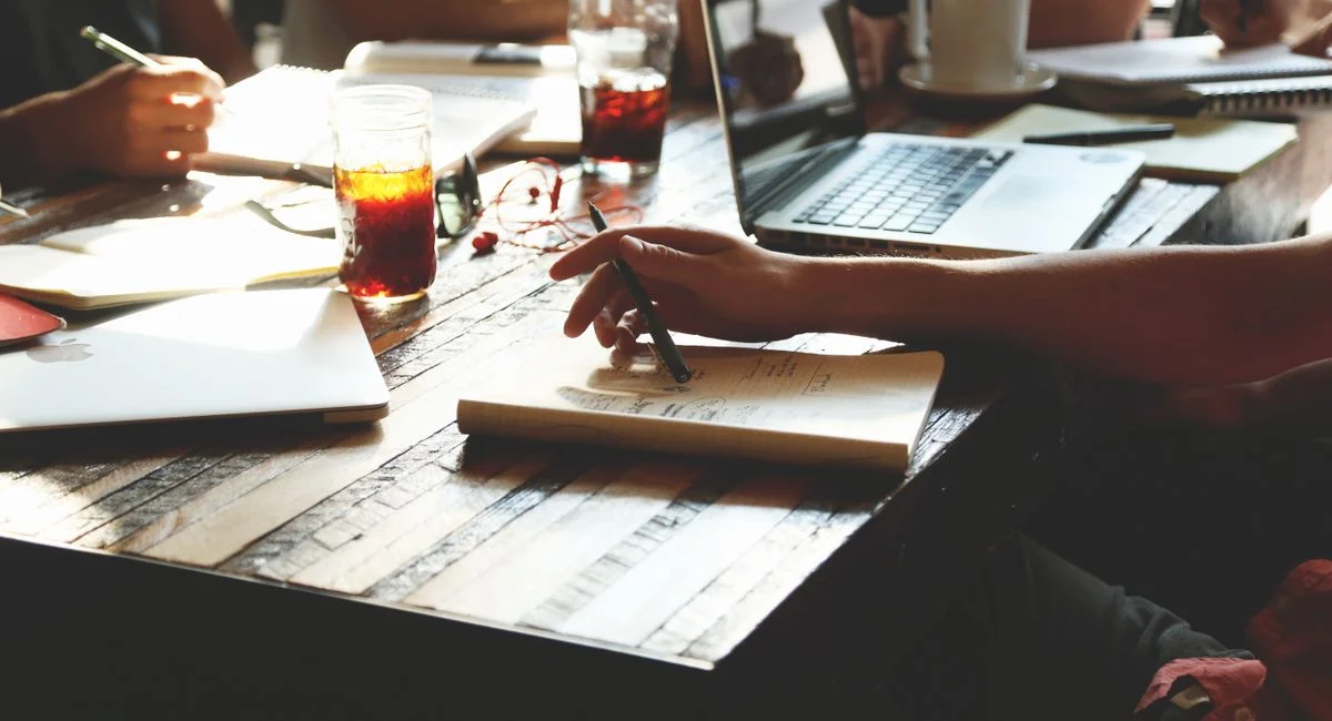 A group of adults engaged in a collaborative office meeting, discussing ideas with notes, laptops, and refreshments.