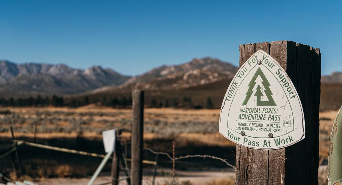 Close-up of National Forest Adventure Pass sign on wooden post with mountains in the background.