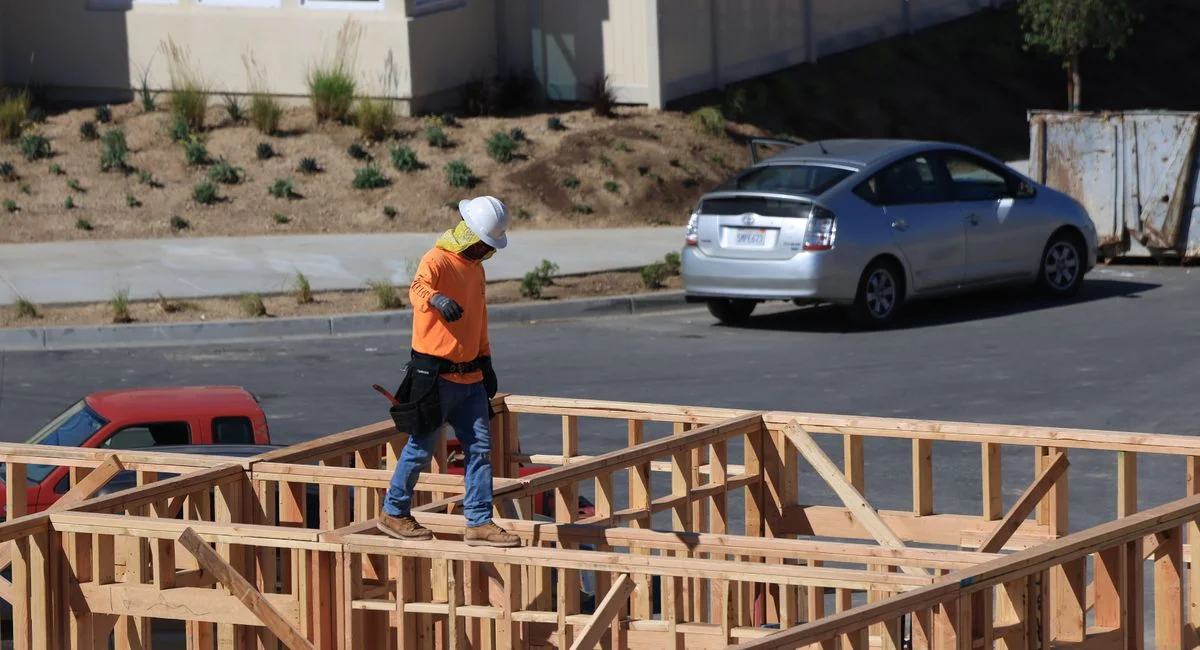 A construction worker wearing safety gear walks on wooden framing at a construction site.