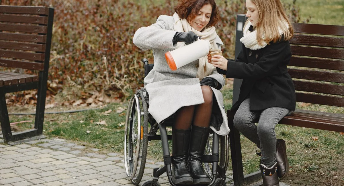 A woman in a wheelchair shares a warm drink with a girl in a park.