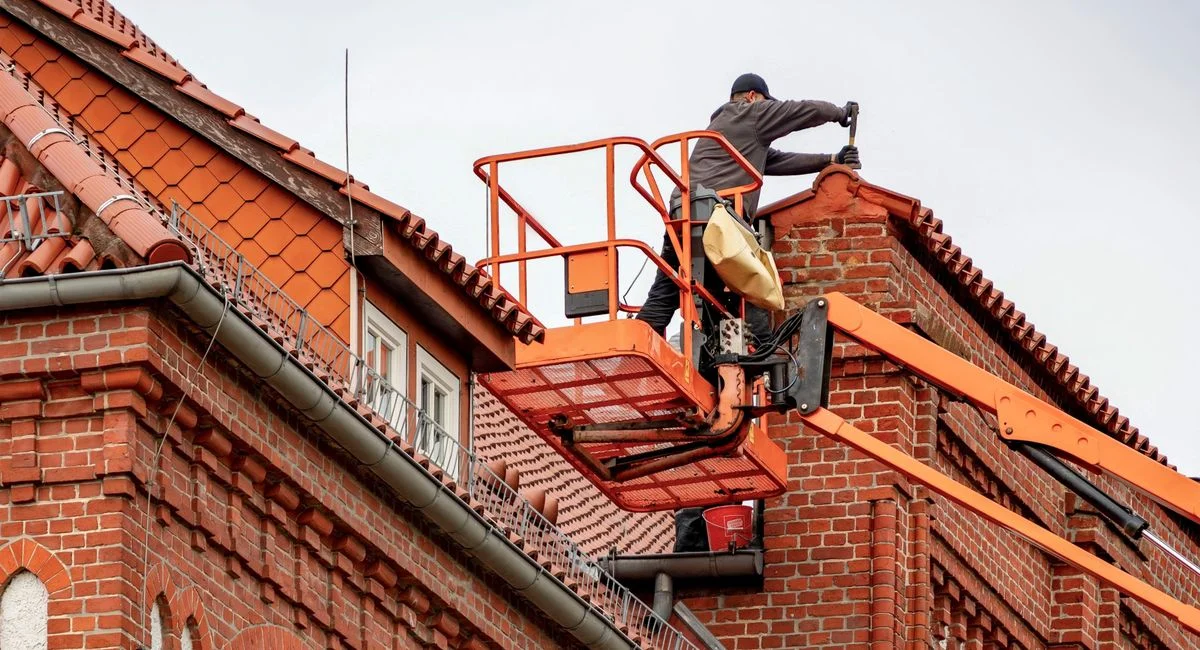 Roofer in a cherry picker conducting maintenance on a red brick building with a tiled roof.