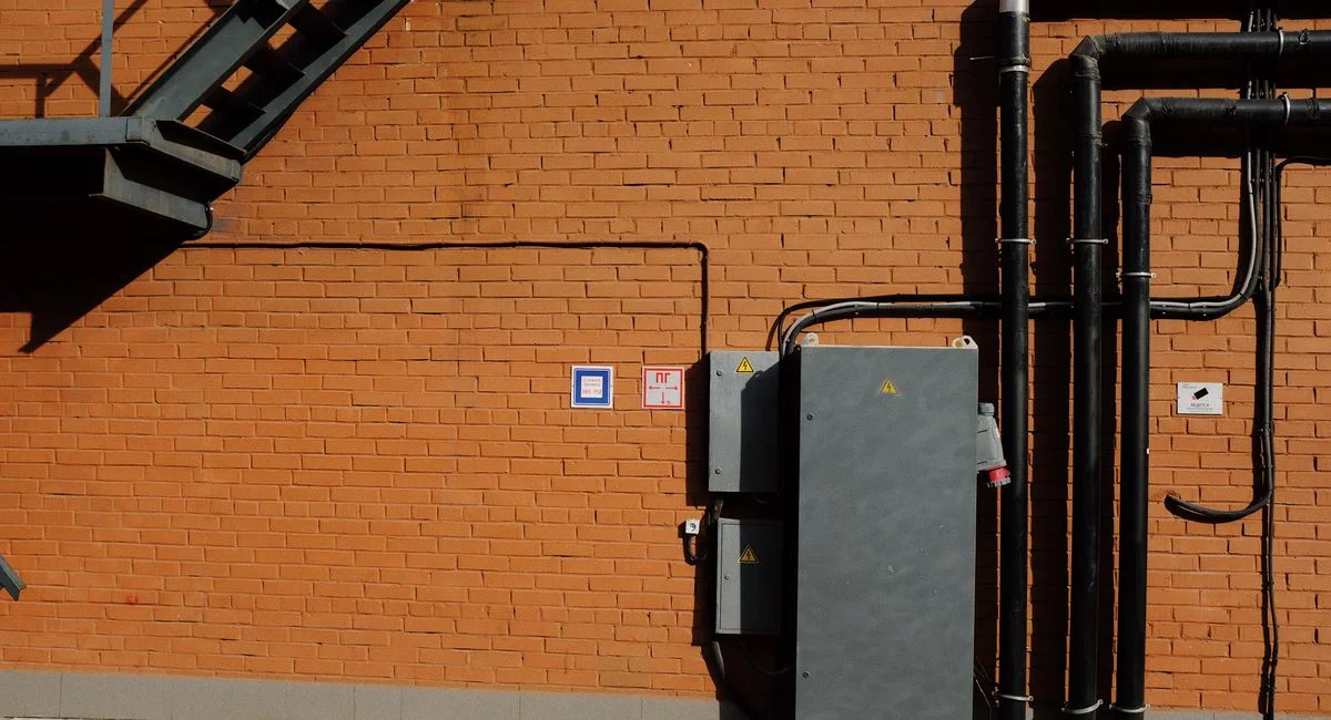 Facade of modern building with metal staircase and transformer box near black metal tubes placed at brick wall